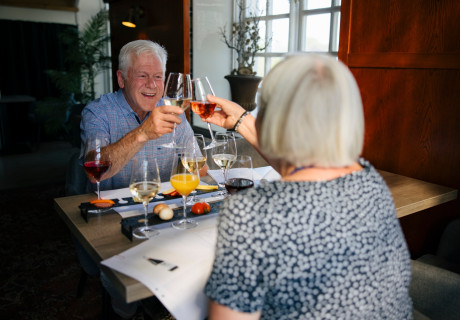 Ett par på dryckesprovning i Eksjö som skålar med glädje i ögonen, inomhus längs med ett fönster.